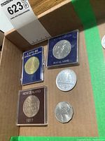 Photo showing six coins and medallions laid in a cardboard box, including Expo 86 official medallion, New Zealand coins in protective cases, Canadian Big Nickel, Australian silver dollar, and a smaller coin.