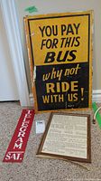 Photo showing all three signs together: large yellow and black bus sign, thin red metal telegram sign, wooden framed paper tender notice.