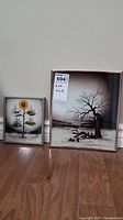 Two framed prints shown side by side on floor. Smaller to left with sunflower image, larger to right with person chopping wood and bare tree.