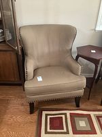 Full front and side view of beige upholstered wing chair with nail head trim and attached seat cushion near wooden furniture and rug.