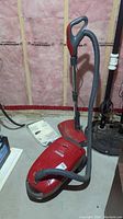 Red Kenmore Elegance vacuum cleaner with hose and floor attachment, placed on a concrete floor near unfinished insulation wall.