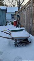 Two wheelbarrows shown outdoors in snow along a fence, one modern green with yellow handles, one vintage light blue with wooden handles.