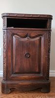 Front view of vintage solid wood stand showing cupboard door with ring pull, open shelf above, and carved details on sides and edges.