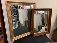 Two framed mirrors side by side on a table against a wall, showing gold frame larger mirror and brown wood frame smaller mirror.