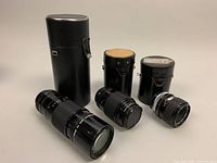 Photo showing all three Canon FD lenses with their respective black cylindrical carrying cases. The 200mm lens is longest and the 50mm is shortest.