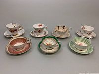 Seven bone china teacup and saucer sets arranged in two rows showing variety of floral and gold-trimmed designs.