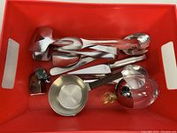 Top-down view inside red bin showing multiple stainless steel utensils and the Alessi Ovolini spice jar with glass and steel components.