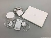White Apple iBook G4 laptop closed with power adapter, cables, and fast wireless charger next to it on table.