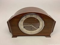 Front view of vintage wooden mantel clock with rounded top and stepped sides, clock face protected by hinged glass cover, visible metal keyhole openings for winding.
