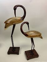 Two crane sculptures displayed side-by-side with wooden bases, both with Lucite bodies and dark wooden heads and necks.