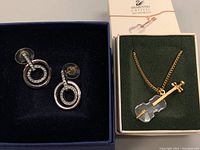 Pair of Swarovski crystal circular earrings with 14K gold posts in navy blue jewelry box next to a gold-tone chain necklace with a violin pendant in a green box.