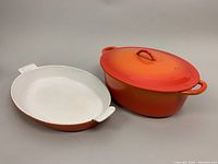 Photo showing au gratin dish with white interior and oval shape along with covered oval casserole dish in flame orange finish.