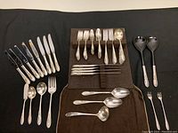 Full layout of all flatware pieces showing forks, knives, spoons, serving utensils and salad tongs spread out on black fabric and flatware roll pouch.