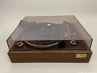 Front view of turntable with closed clear dust cover, showing wood finish base and controls.