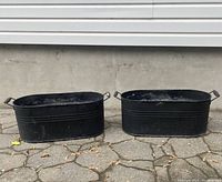 Pair of black galvanized metal tubs shown side by side on stone pavement outside, with visible wear.