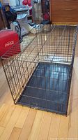 Large wire dog crate set up with black plastic tray inside on wood floor.
