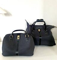 Two black handbags with leather handles and gold logo plaque on front, one carry-on and one wheeled glider suitcase.