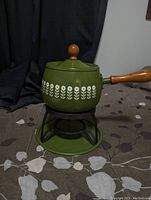 Front-side angle of green enamel fondue pot on matching metal stand showing floral motif and wooden handle and knob.