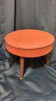 Photo showing front angle of a round orange upholstered foot stool with wooden legs and brass tipped feet.