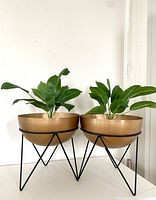 Pair of large metal planters with bronze bowls and black hairpin legs, each containing a green faux plant, placed side by side against a white background.