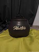 Front angle of the matte black Planters cauldron-style container showing embossed logo and two small side handles.