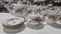 Complete view of floral china tea set with teapot, sugar bowl, creamer, cups, and saucers, featuring pink roses and gold trim.
