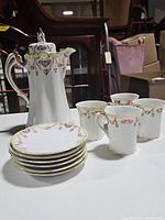 Photo showing tall coffee pot with lid, four coffee cups, and six small plates stacked
