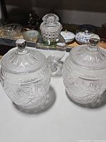 Three clear antique glass canisters shown from above angle, highlighting etched details and glass lids with knobs.