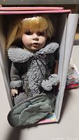 Porcelain doll with blonde hair, grey sherpa jacket with pom-poms, tucked in blue-green pants inside its box.