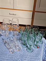 Assorted group of stemmed clear wine glasses, green goblets, and small glass jars on a blue tablecloth.