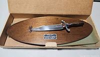 Overall view of the Bowie knife mounted on an oval wooden plaque inside its original box.