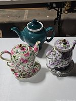 Photo showing three teapots including a round teal ceramic teapot and two floral ceramic teapots each with matching teacup and saucer.