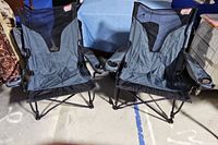 Two gray Coleman folding camping chairs with mesh backs side-by-side, showing cup holders in right armrests and their black folding metal frames visible beneath.