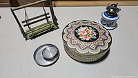 Four assorted decorative housewares on table: green swing, floral box, silver tray with coasters, ceramic table lighter.