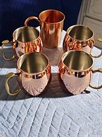 Five copper mugs arranged in a group, four round with brass handles, one taller and cylindrical without a brass handle.