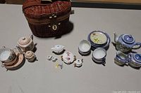 Overview photo showing four children's tea sets arranged on a table with a woven basket, cups, teapots, sugar and cream containers, and small plates.