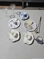 All eight candlestick holders and candle snuffer shown from a top view on a flat surface, various ceramic with floral designs and two clear glass holders visible.