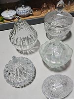 Top angled view showing five different vintage clear glass candy jars with intricate textures and lids on a white surface.