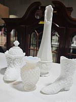 Four assorted vintage milk glass pieces placed on white surface with dark furniture background: tall vase, lidded jar, small vase, boot-shaped vase.
