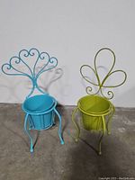 Both blue and green vintage metal plant stands shown side by side, blue has a scrolled heart-shaped back, green has a clover-shaped back with pot