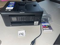 Black Brother all-in-one printer with attached power cord, ink cartridge box, and a separate cartridge placed next to the printer on a table.