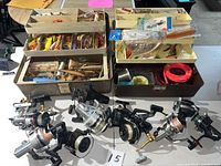 Photo showing 11 fishing reels and two open tackle boxes with assorted lures, line, hooks, and fishing accessories on table.