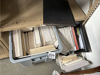 Plastic bin filled with stacked white mat boards, foam boards, and some wooden framing strips under a shelf.