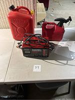 Three items on a table: two red gasoline cans (one large, one small) and a Century Battery Charger with cables.