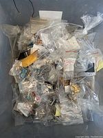 A large bin filled with multiple small clear plastic bags containing various pieces of raw jewelry findings, costume jewelry, charms, earrings, and hair barrettes. Items show a range of metallic finishes and colored stones or enamel.