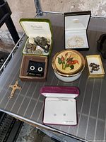 Overview of assorted jewelry and vanity items including pearl bracelet, green box of buttons/coins, black pearl earrings, crystal earrings, crucifix, and decorative round powder jar.