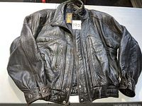 Brown leather bomber jacket laid flat on table, showing front with zippered pockets and snap cuffs.