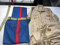 Photo showing folded navy blue pants with red vertical stripe and beige belt alongside folded beige khaki shirt with ribbons and additional beige pants and belt.