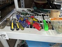 Assortment of various hand tools including screwdrivers with colored plastic handles, sledgehammers, ratchet set parts, and a spool of twine on a white grate surface.