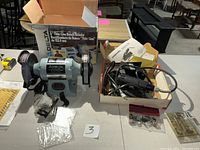 Delta 6-inch Thin Line Bench Grinder, new in box with manual and accessories, alongside a box containing a used Dremel Moto-Flex tool and accessories.